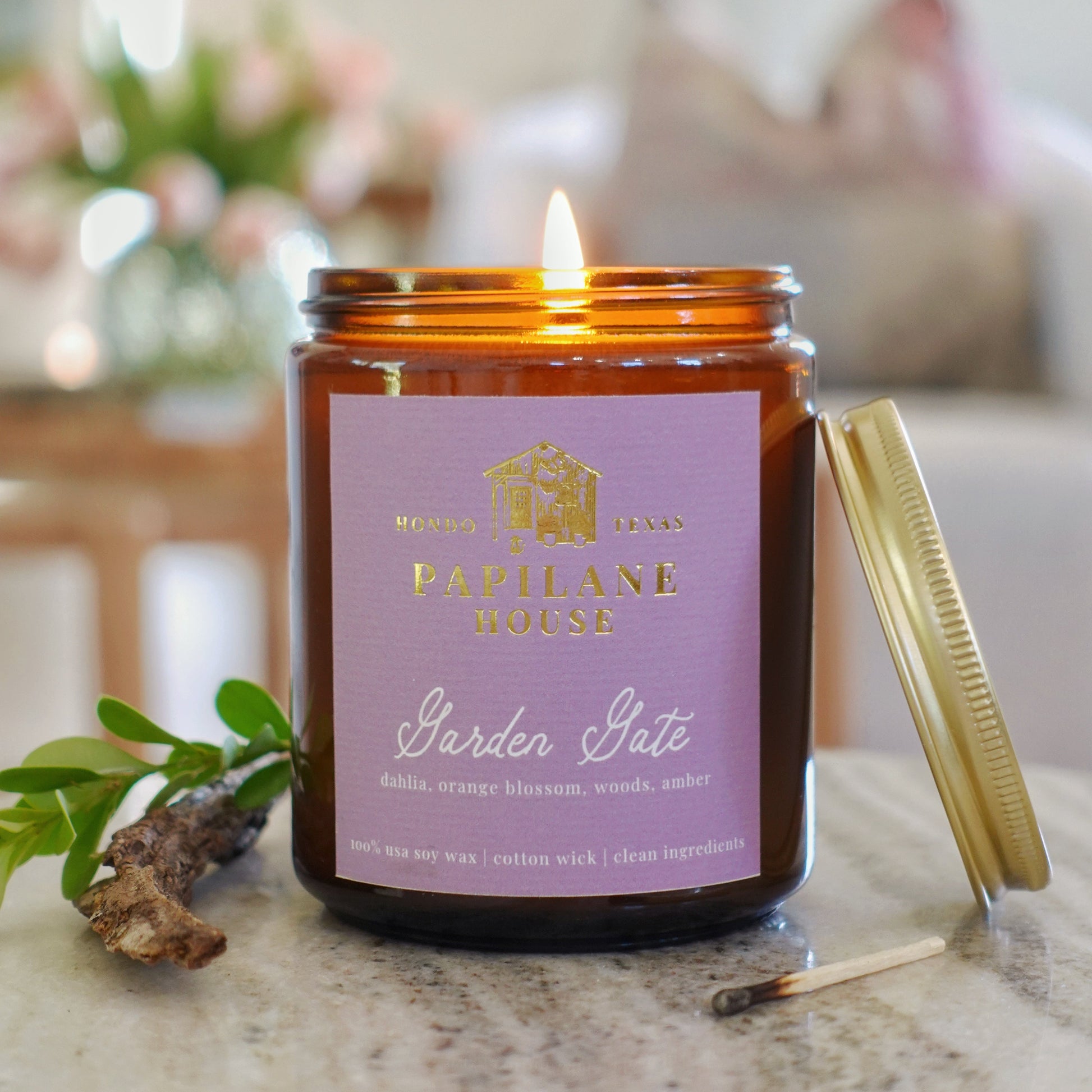 An amber glass jar candle with a lit flame sits on a stone surface. The candle has a lavender label reading 'Papilane House Garden Gate' with gold text, and a gold-toned lid rests beside it.