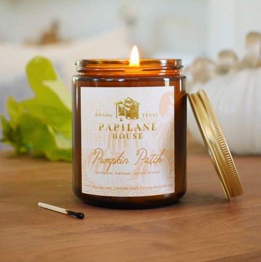 A lit amber glass jar candle with a gold lid resting beside it on a wooden surface. The front label reads 'Papelane House' and 'Pumpkin Patch,' with a small match placed nearby.