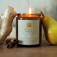 A lit amber glass jar candle with a light green label reading Papilane House Pear Tree Lane, styled on a wooden surface with ginger root, cinnamon sticks, and a pear in the background.