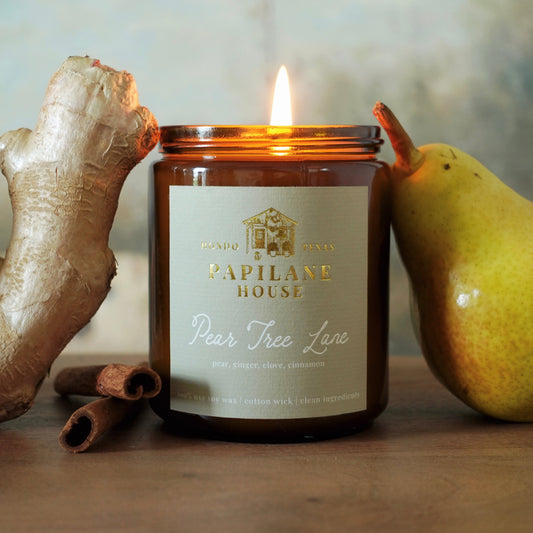 A lit amber glass jar candle with a light green label reading Papilane House Pear Tree Lane, styled on a wooden surface with ginger root, cinnamon sticks, and a pear in the background.