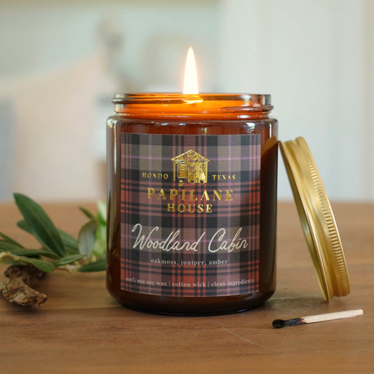 A lit amber glass jar candle with a gold lid resting beside it on a wooden surface. The front label reads 'PAPILANE HOUSE' and 'Woodland Cabin' with a plaid pattern.